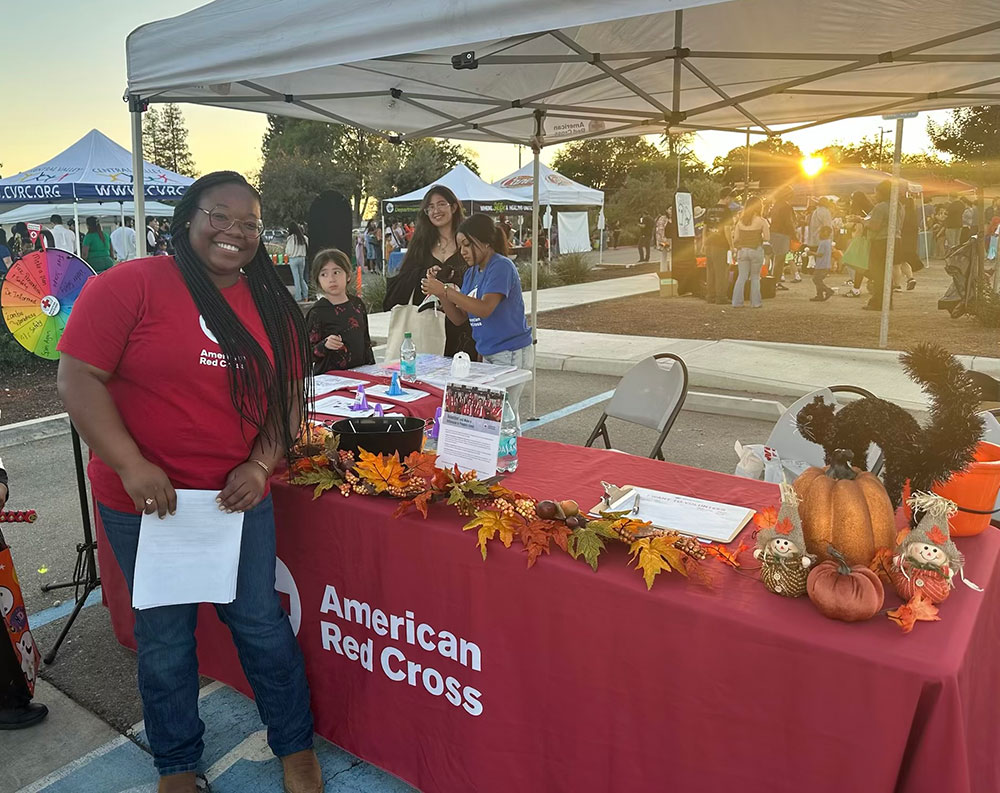 A Red Cross volunteer working at a Red Cross booth at an event held in a parking lot.