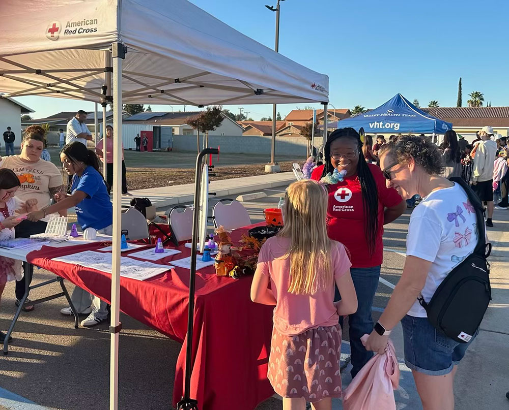 a Red Cross volunteer speaking with a young child and adult at a Red Cross booth at an event in a parking lot.