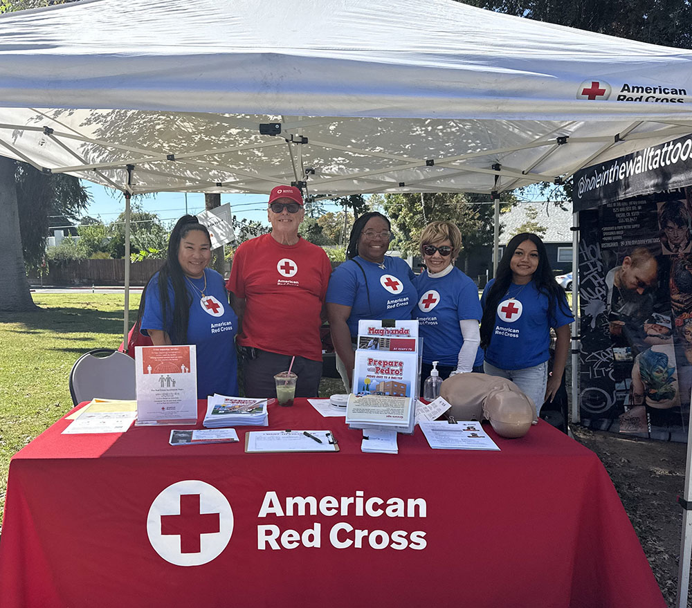 Four Red Cross volunteers smiling and standing at a Red Cross booth at an outdoors event.