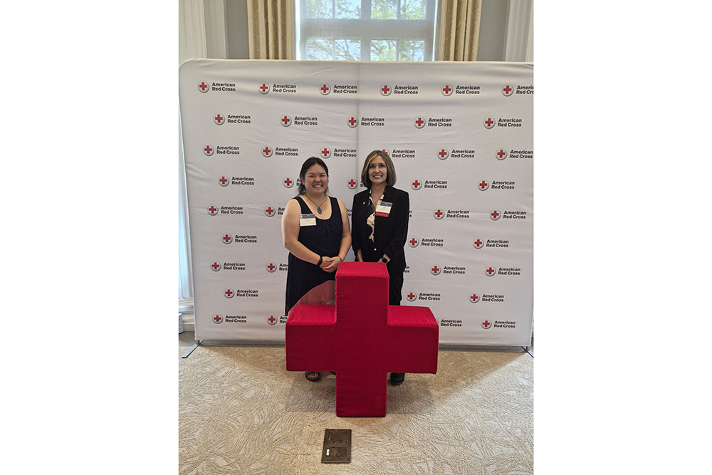 Jody Kimbrel and another person standing behind a large Red Cross sign with a Red Cross backdrop behind them.