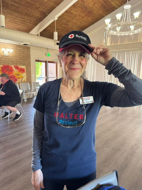 Julie Atwood wearing a Red Cross hat and Halter Project shirt poses in a room, surrounded by soft lighting and neutral-colored walls.