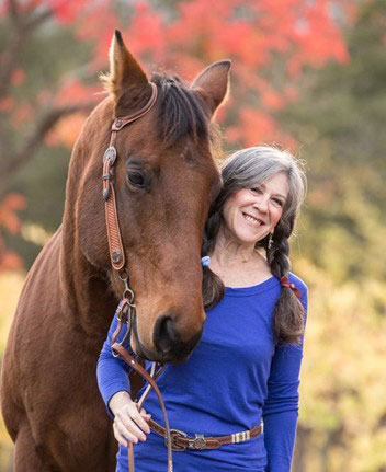 Julie Atwood in a blue shirt stands beside a brown horse in a natural outdoor setting.