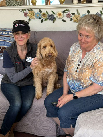 Julie Atwood and another woman sit on a couch, smiling, with a dog resting comfortably between them.