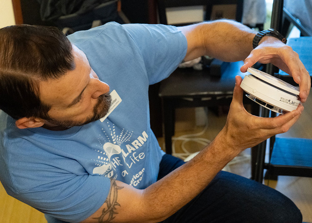 Justin Mueller in a blue shirt holds a smoke alarm. He is focused, carefully holding and examining the device.