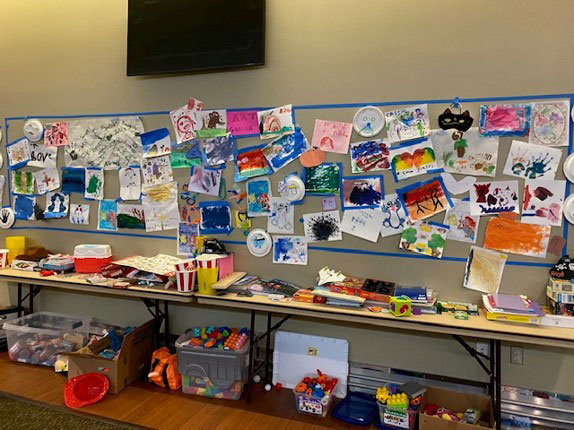 children's drawings and paintings pinned on a large board. Below, a table holds various art supplies and toys.