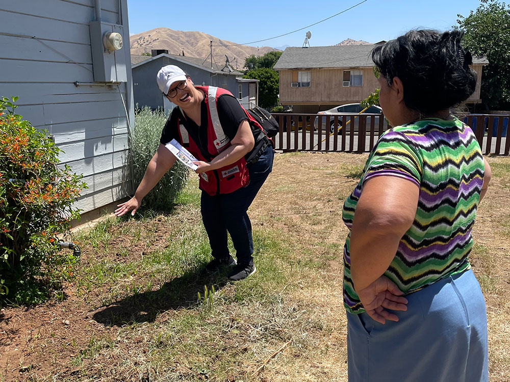 Margarita Moreno, wearing a Red Cross vest and hat, talking to a resident next to the side of a house.