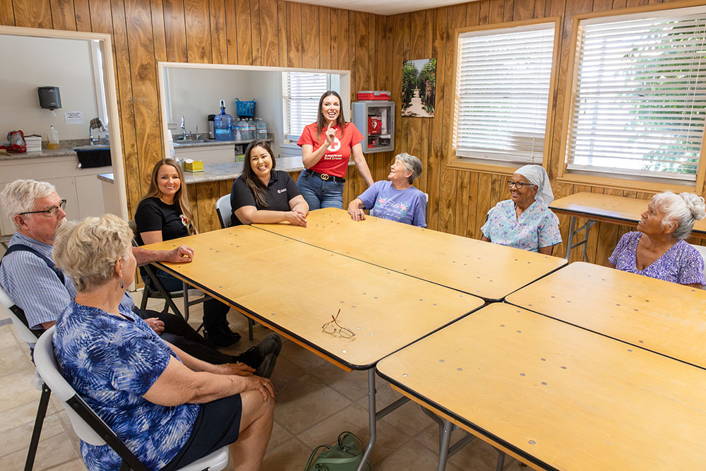 Margarita Moreno and two other Red Cross volunteers speak to a group of seniors inside a room with folding tables.