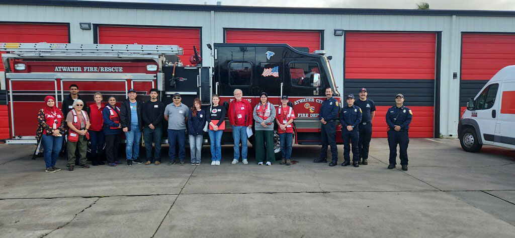 A group of Red Cross and firefighter volunteers stand in front of a fire truck labeled "Atwater Fire/Rescue." Red garage doors and a fire station building form the backdrop.