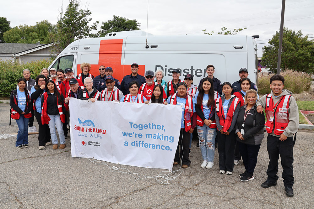A group of diverse Red Cross volunteers in red vests stand in front of an emergency response vehicle, holding a sound the alarm banner reading, "Together, we're making a difference."