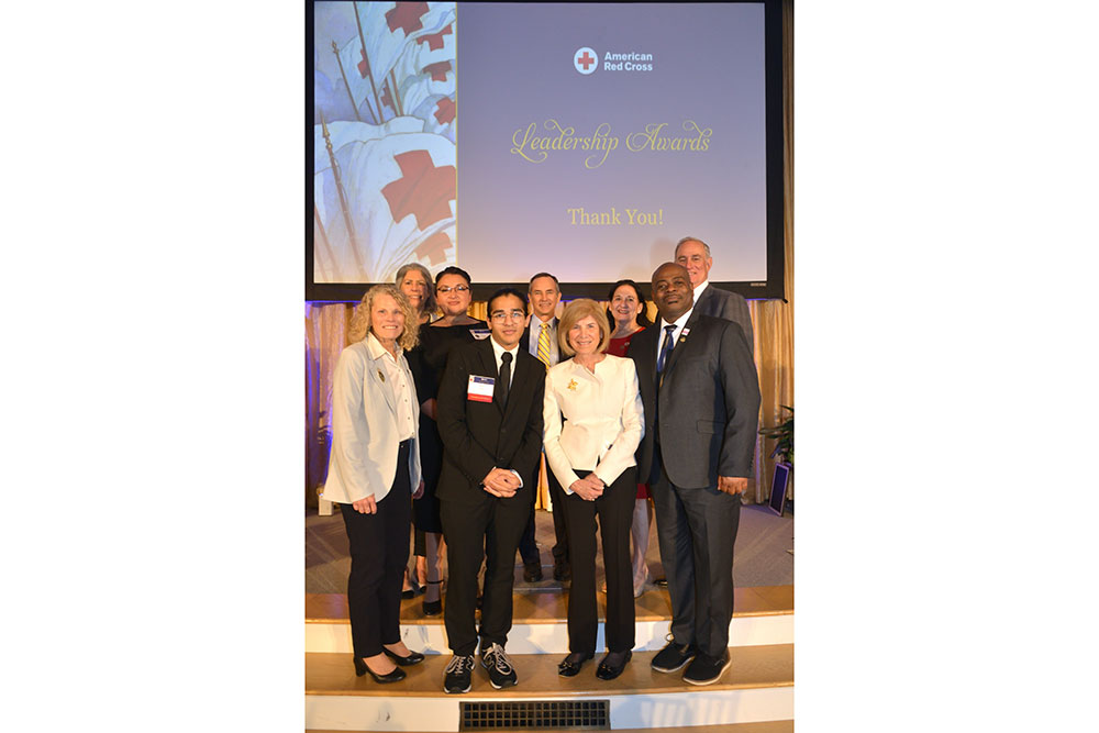 Razi Aftab and eight other people, in dress clothing, with five people standing on a stage and four on stairs, at a Red Cross event.