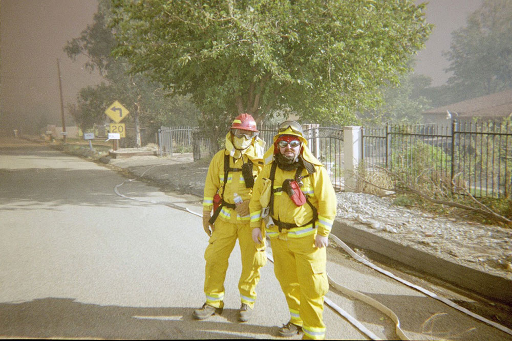 two firefighters in safety gear on a street with smoke and trees in the background.