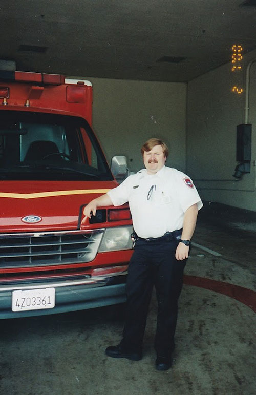 Steven Bradley wearing an EMT uniform poses for picture while standing next to an ambulance.