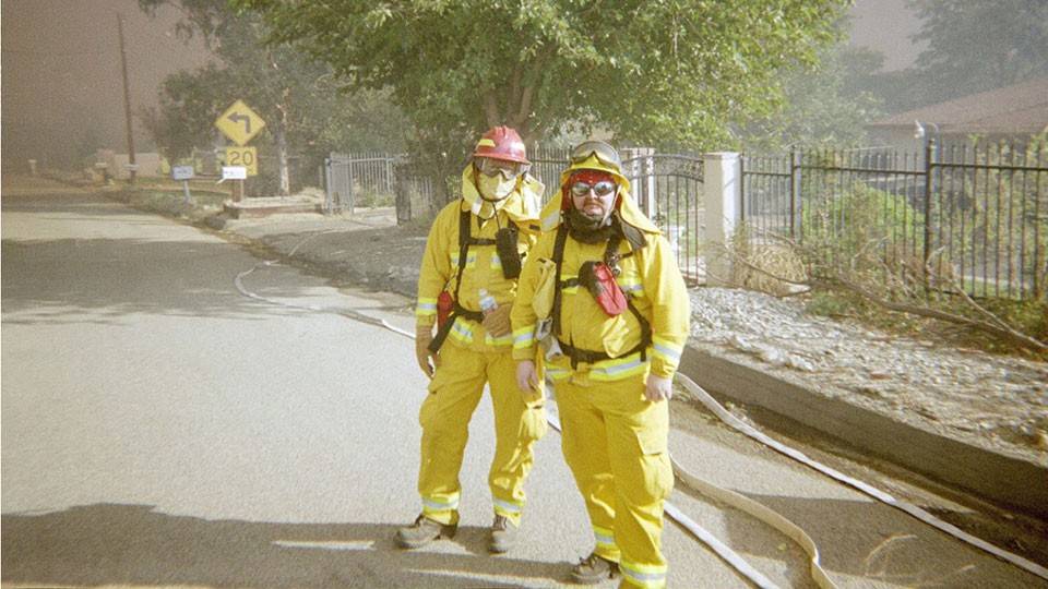 two firefighters in safety gear on a street with smoke and trees in the background.