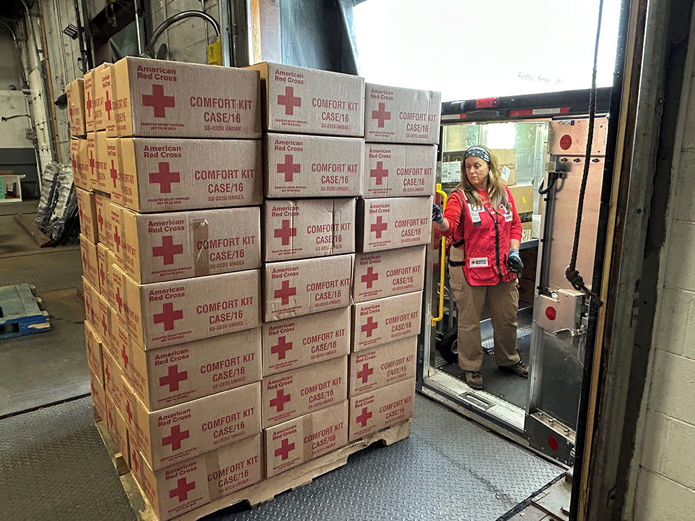 Red Cross boxes on a pallet sitting on a loading dock with a Red Crosser checking them.