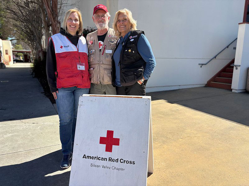 Three smiling Red Cross volunteers stand outside, wearing branded vests and badges, near a sign for the Silicon Valley Chapter. Sunny day.