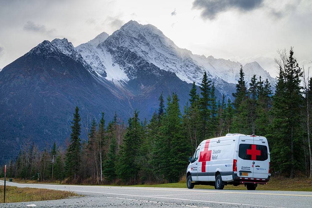 Red Cross Disaster Relief van on the road next to tall trees and a snowy mountain.