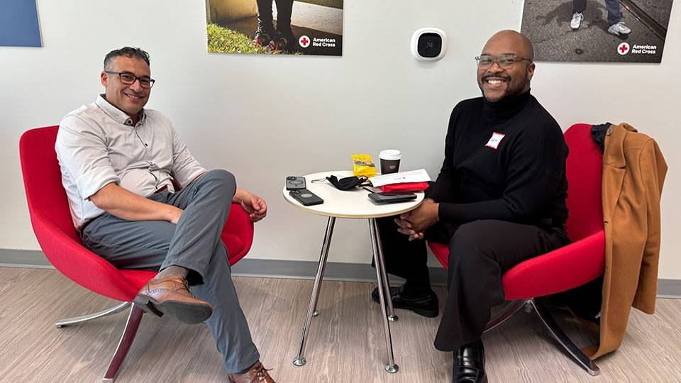 Ed Faso and Noah James sitting in chairs with a small round table between them, at the Red Cross blood laboratory in San Leandro