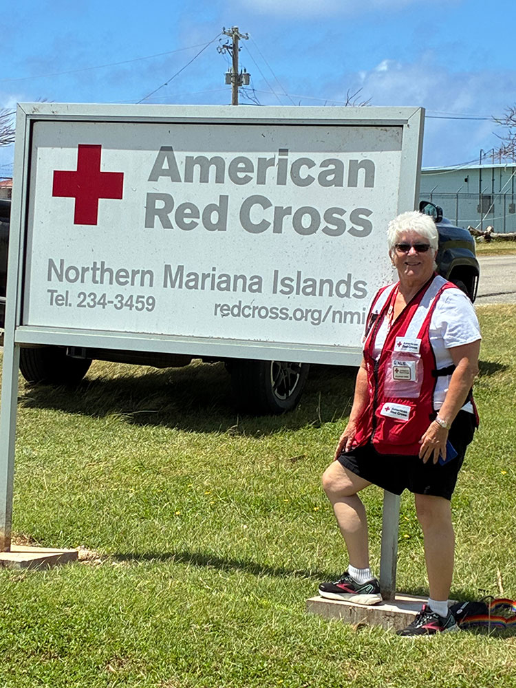 Red Cross volunteer Gale Carli in Saipan standing on the grass in front of the American Red Cross Northern Mariana Islands sign.