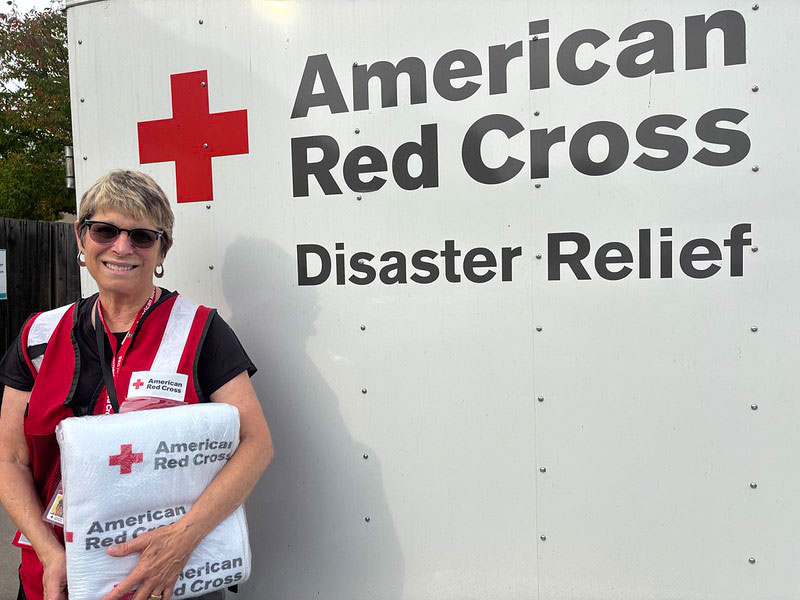 Volunteer Leslie Grant holding a wrapped Red Cross blanket and standing in front of a Red Cross Disaster Relief trailer.