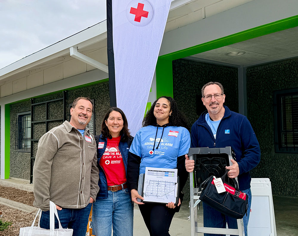 Four smiling volunteers stand outside a building with a Red Cross banner. They're wearing casual clothing and holding equipment.