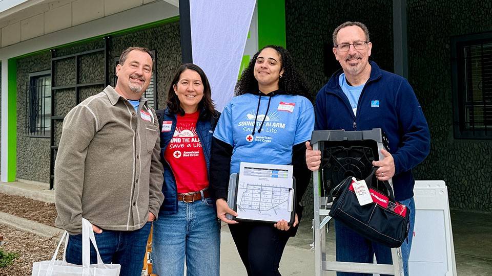 Four smiling volunteers stand outside a building with a Red Cross banner. They're wearing casual clothing and holding equipment.