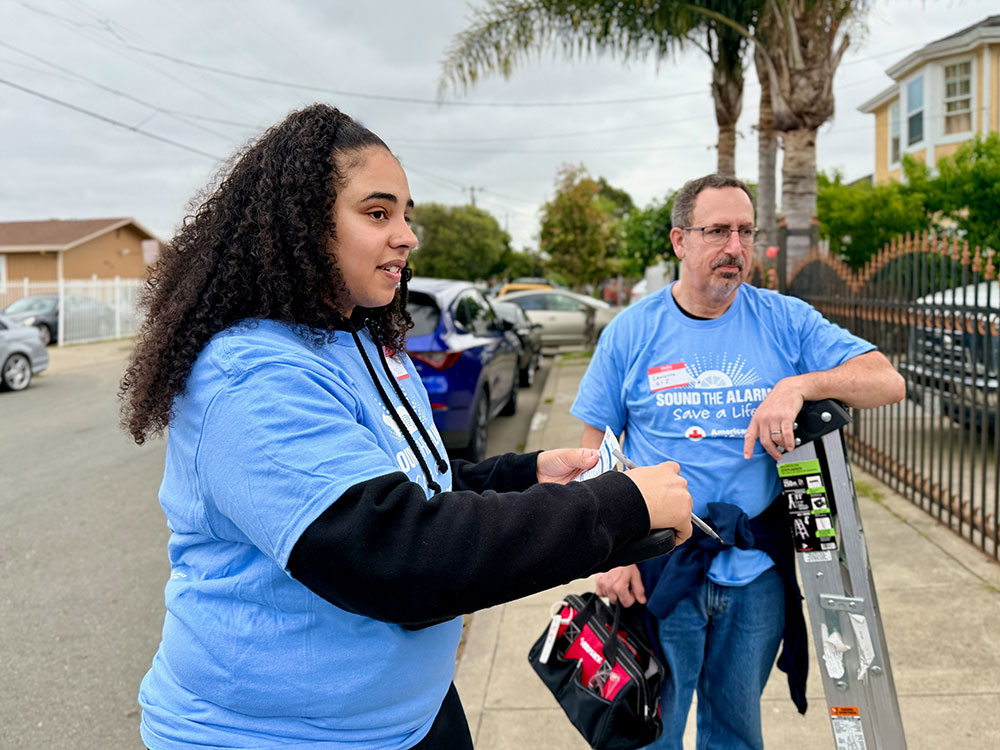 Tierra and Laurence wearing light blue shirts labeled "Sound the Alarm, Save a Life" stand on a sidewalk. One holds a flyer, the other a ladder.
