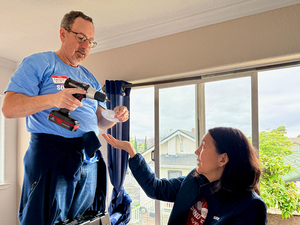 Cymbre passes screws to Laurence as he installs a smoke alarm. Bright setting with visible trees outside.
