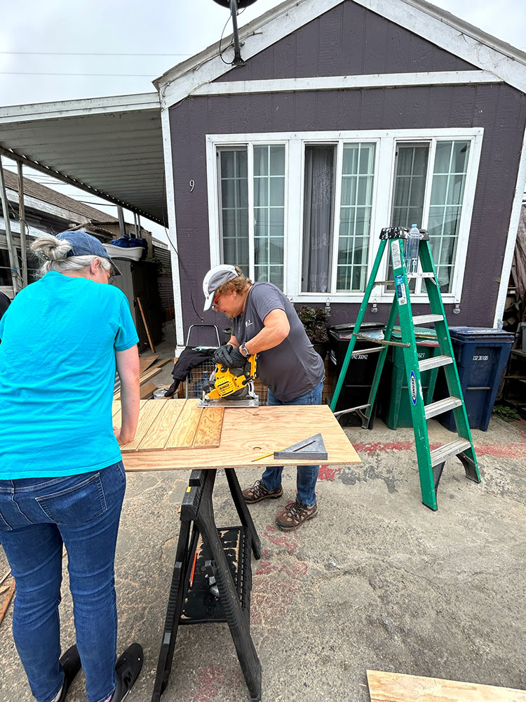 two volunteers cutting wood on a sawhorse in a backyard.