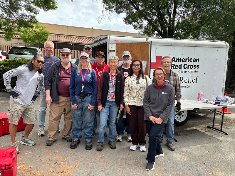 A diverse group of Red Cross volunteers pose smiling in front of a Red Cross truck.