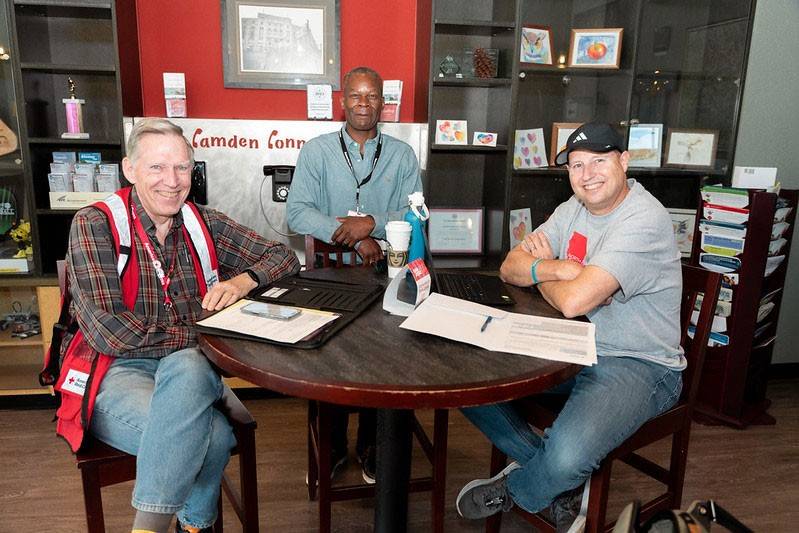 Three men sit at a round table in a cozy room, engaged and smiling. One wears a Red Cross vest. Laptops, papers, and a coffee cup are on the table.
