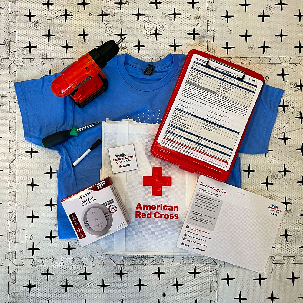 Emergency preparedness kit on patterned surface, featuring a blue T-shirt, red drill, smoke detector, screwdriver, clipboard with forms, and American Red Cross tote bag.