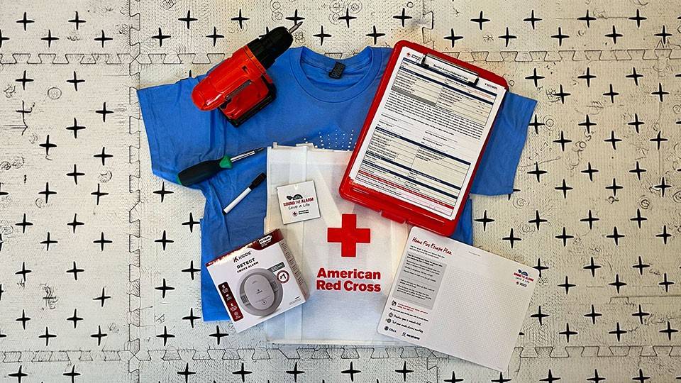 Emergency preparedness kit on patterned surface, featuring a blue T-shirt, red drill, smoke detector, screwdriver, clipboard with forms, and American Red Cross tote bag.