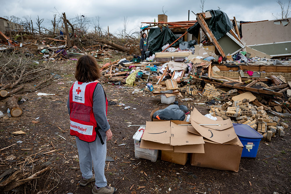 Red Cross volunteer standing next to destroyed house with debris all around.