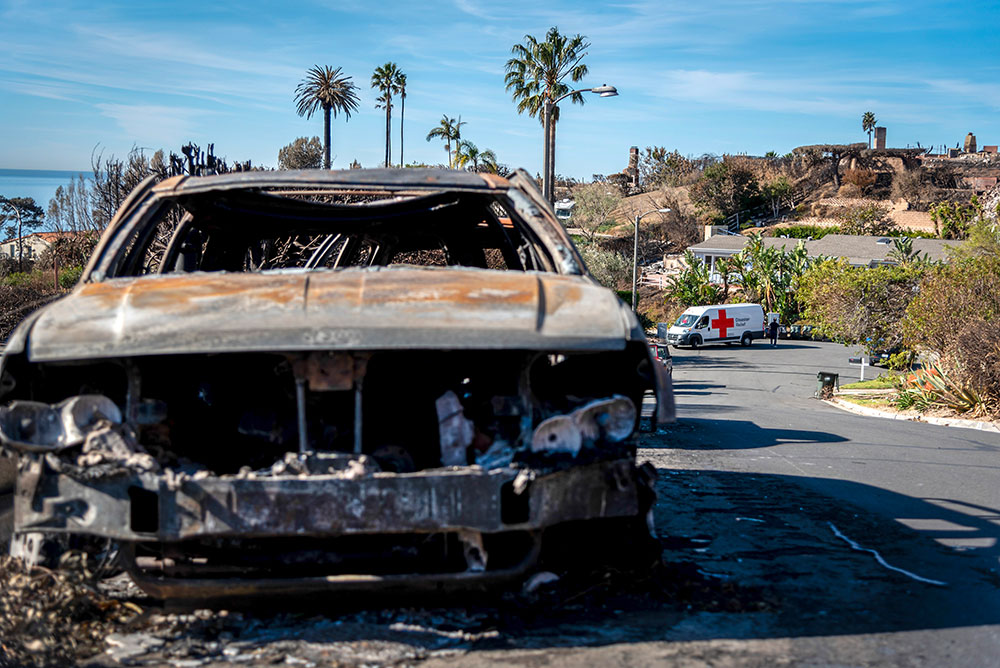 burned car on the side of the road with a Red Cross Disaster Relief vehicle in the background..