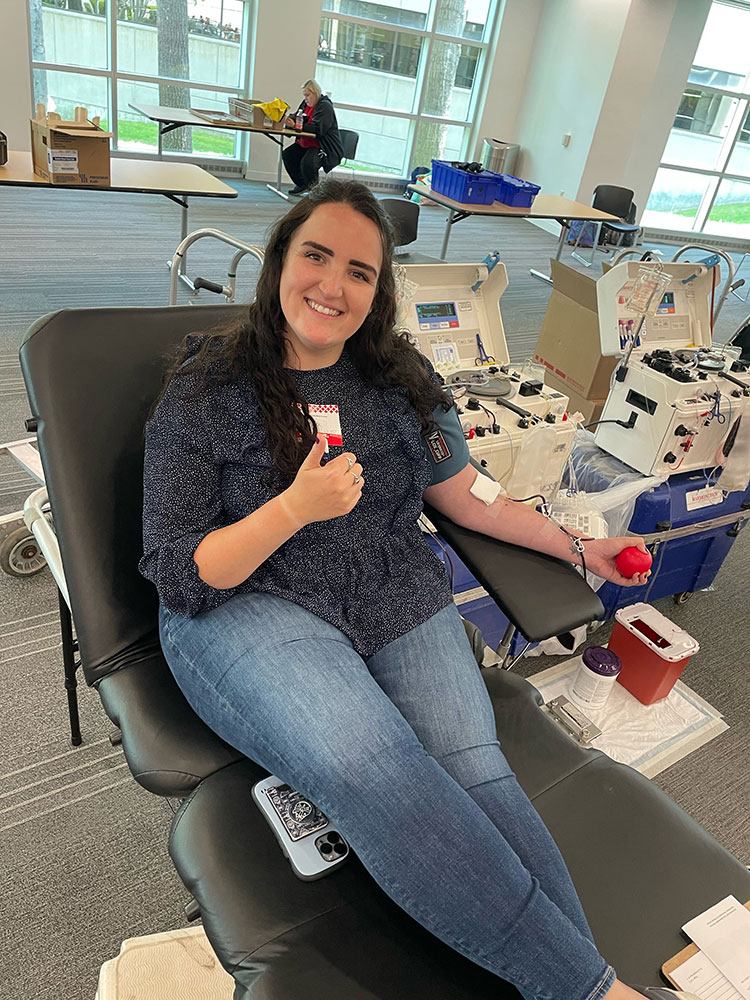 A woman smiling and giving a thumbs-up while donating blood. She is seated on a recliner, holding a red stress ball. Medical equipment is visible around her.