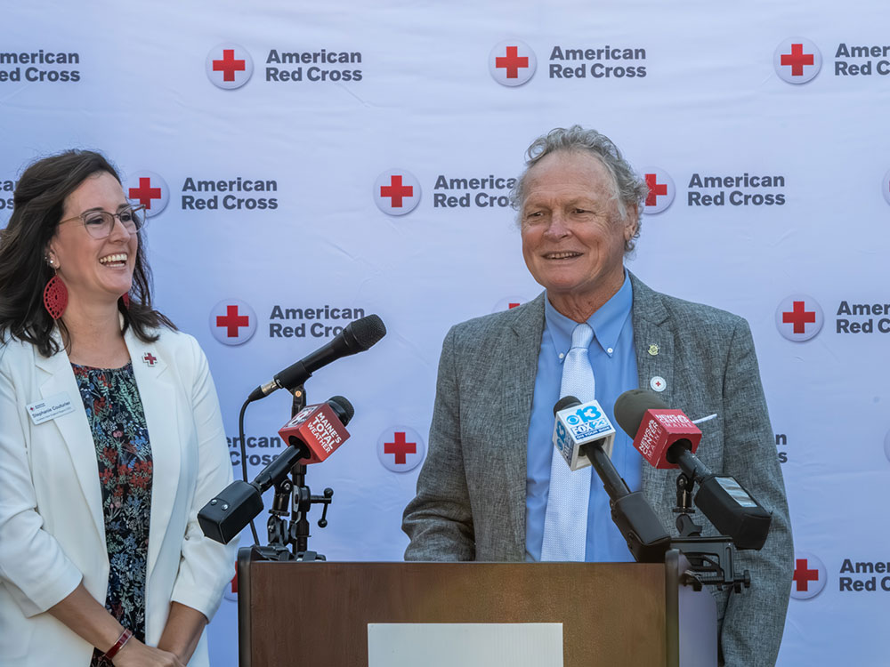 two people at podium with one speaking on a microphone, with red cross logos in the background.