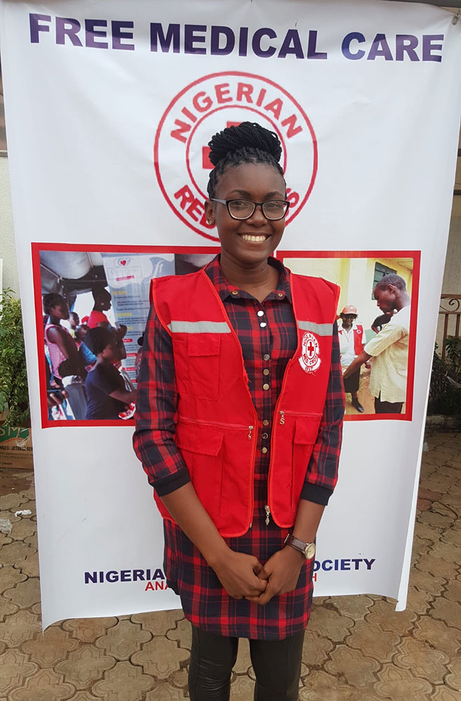 Red Cross volunteer Chinenye Angela Okoli standing in front of a Red Cross Free Medical Care banner.