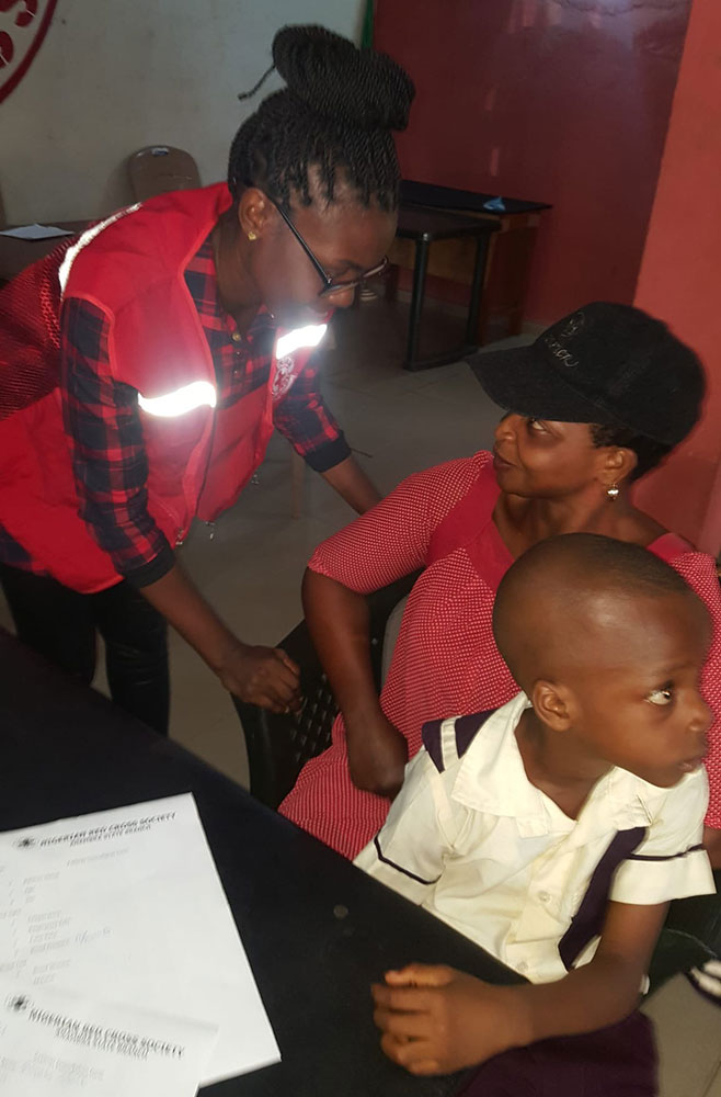 Red Cross volunteer Chinenye Angela Okoli standing next to a mother holding her child.