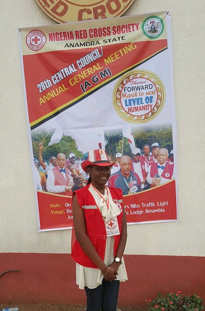 Red Cross volunteer Chinenye Angela Okoli standing in front of a Red Cross 28th Central Council Annual General Meeting banner.
