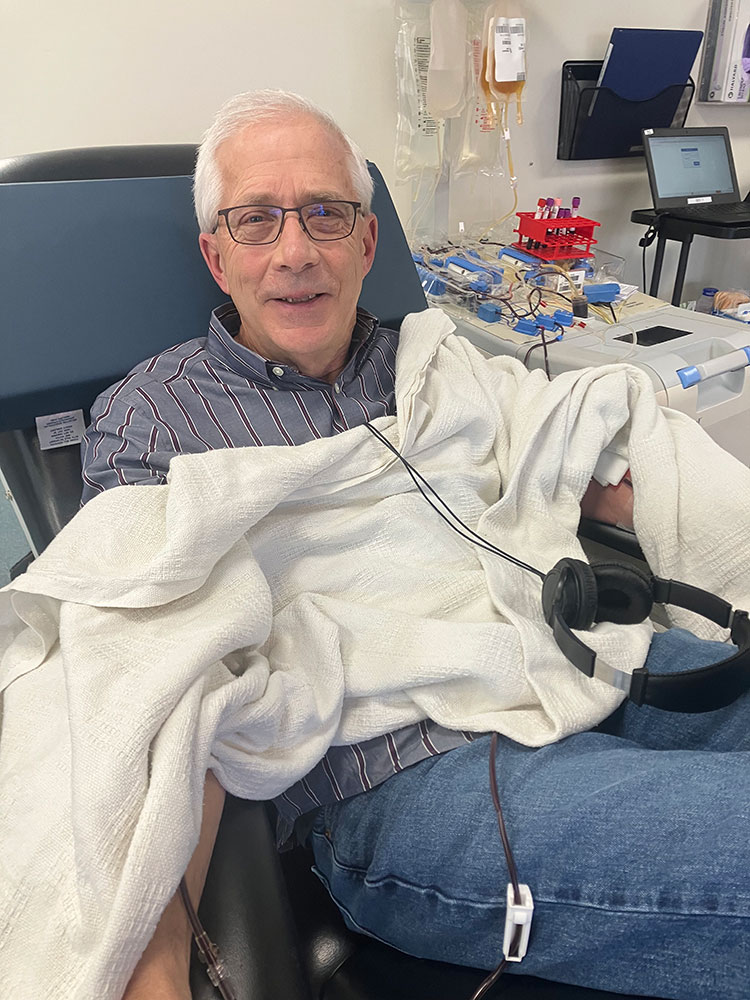 Brad Eldred wearing glasses, heaphones and a small blanket, sitting in chair at blood donation center and donating blood.