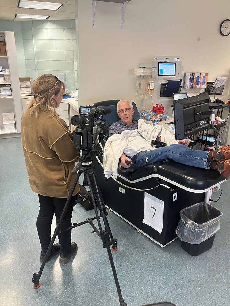 Brad Eldred wearing glasses, heaphones and a small blanket, sitting in chair at blood donation center and donating blood while a person with camera interviews Brad.