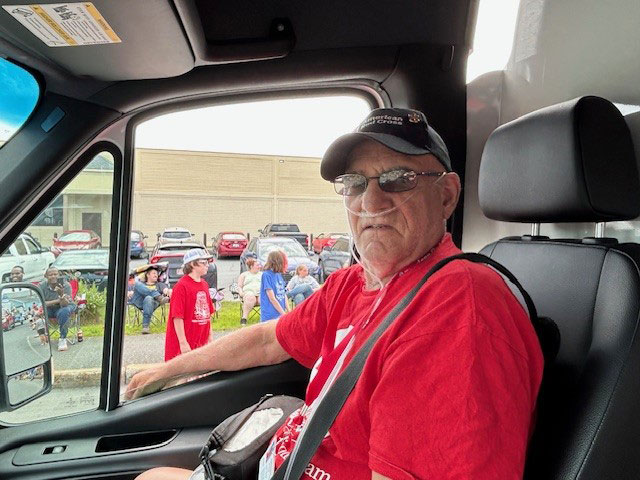 Bill Thomas, wearing a red cross hat and shirt, sitting in a vehicle with adults and childrean outside.