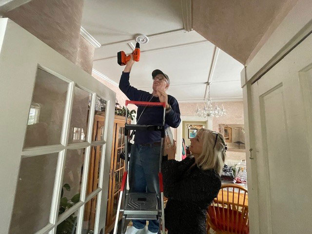 Bill Thomas, standing on a ladder with another person holding it, installs a smoke alarm on a ceiling in a kitchen.