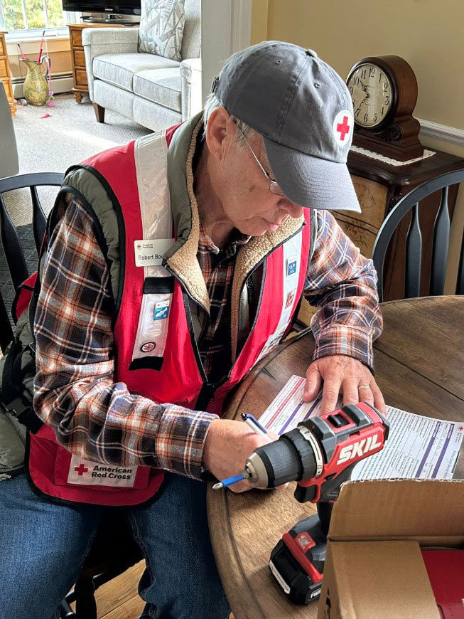 Bob Bouchard in an American Red Cross vest and cap writes on a form at a table with a cordless drill nearby.