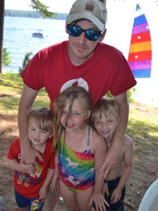 Brian Mayhew in a red cross shirt and cap, smiling with three young children near a lake. The children wear colorful swimsuits.