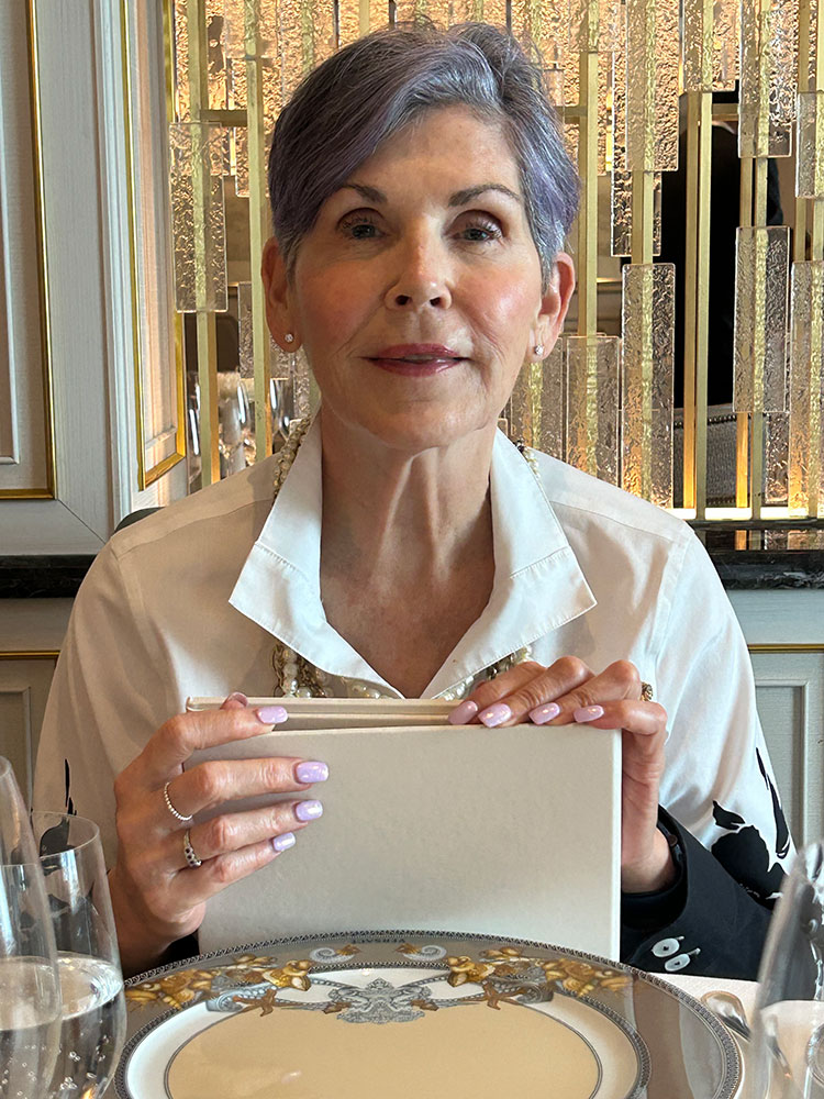 Christine Burke with short gray hair holds a menu at an elegant restaurant. She wears a white shirt; table set with ornate plate and glassware.