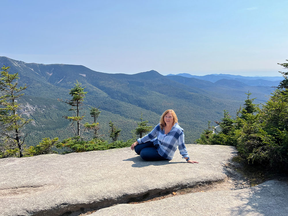 Deb Camejo in a plaid shirt sits smiling on a rocky ledge overlooking expansive, sunlit mountains and trees.