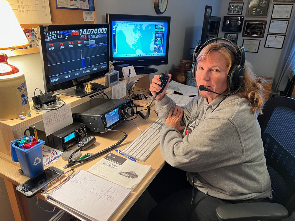 Deb Camejo in a grey sweatshirt operates a radio transmitter at a desk. She wears a headset and holds a microphone. Monitors display frequency data and a map.