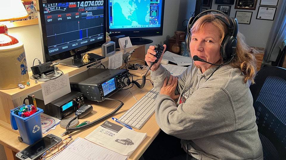 Deb Camejo in a grey sweatshirt operates a radio transmitter at a desk. She wears a headset and holds a microphone. Monitors display frequency data and a map.