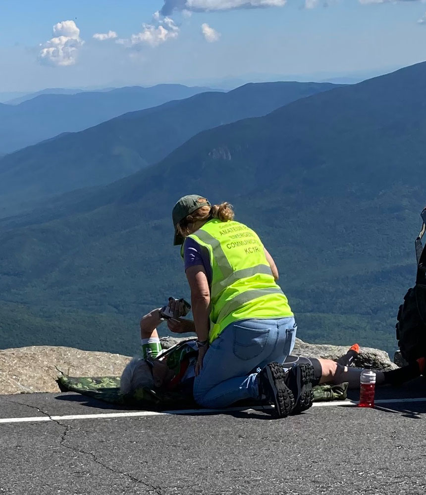 Deb Camejo in a neon yellow vest attends to another person lying on a mountain road, with vast scenic views and blue mountains in the background.